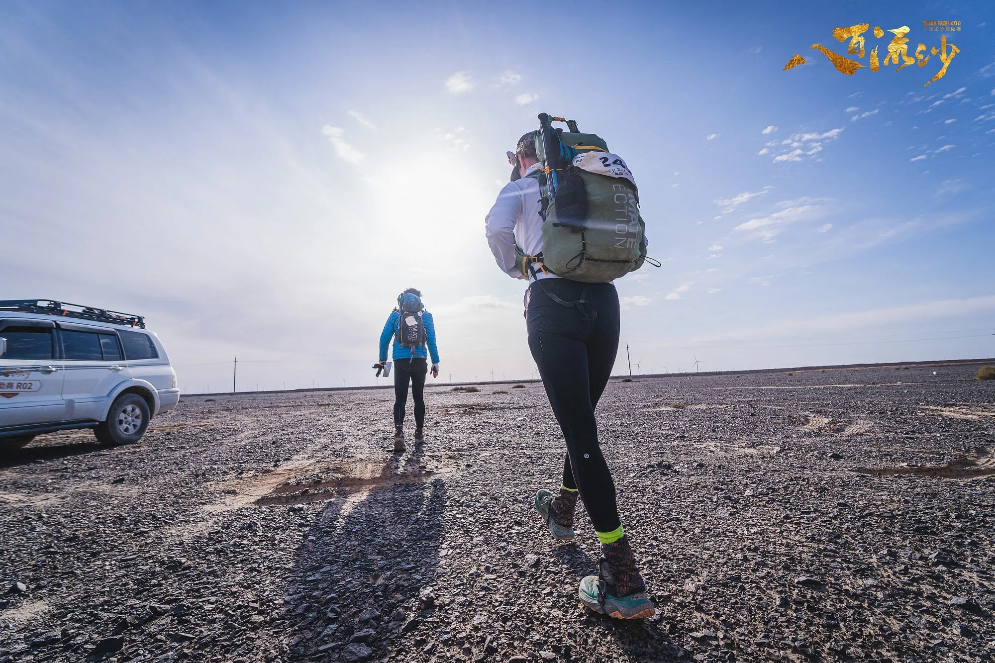 A runner with a race pack silhouetted against a low sun on a wide gravel track in the Gobi.